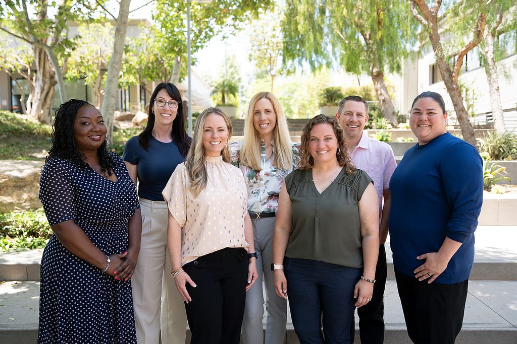 Group of seven members of the ARM team posing and smiling at a business campus surrounded by buildings and trees.