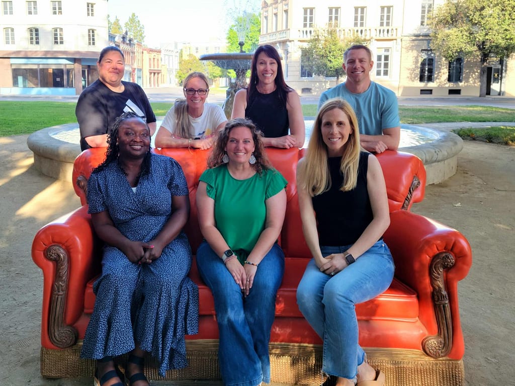 Group of seven members of the ARM team posing and smiling while seated and standing around a large red decorative couch in an outdoor plaza with historic-style buildings in the background.