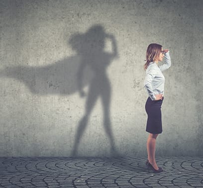 A profile shot of a professionally dressed woman looking into the distance with a hand on her hip. On the wall behind her, the shadow takes the form of a female superhero in a similar pose.