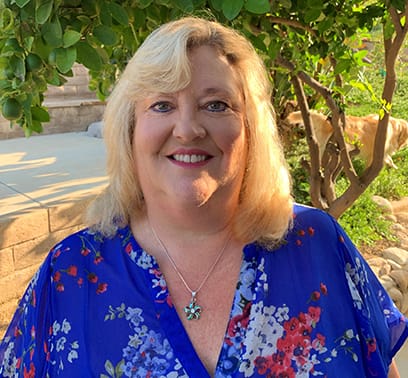 Kim Goldberg is smiling, wearing a floral blouse, and standing in front of a tree.