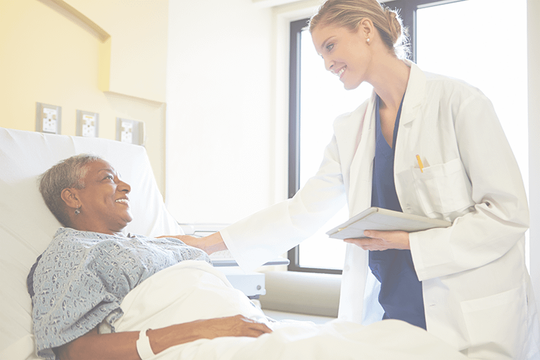 A female hospital patient smiles as she looks up at a doctor holding a tablet. The doctor is resting her other hand on the patient's shoulder.