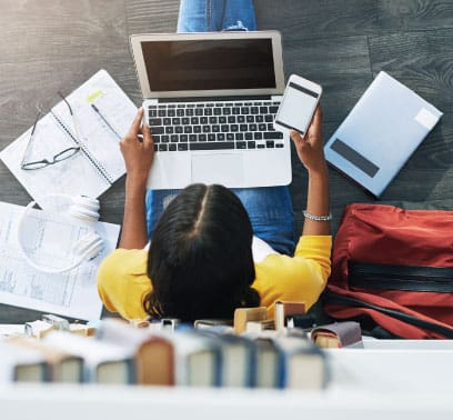A student sits against a bookcase as she browses her mobile phone with an open laptop on her lap. On the floor around her, there is a paper, pair of headphones, notebook, eyeglasses, pen, book, and backpack.