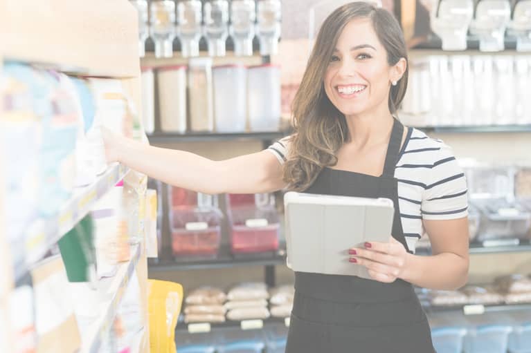 A woman in a striped shirt and black apron smiles for the camera as she checks the store's inventory with a tablet that has mobile floor stock software.