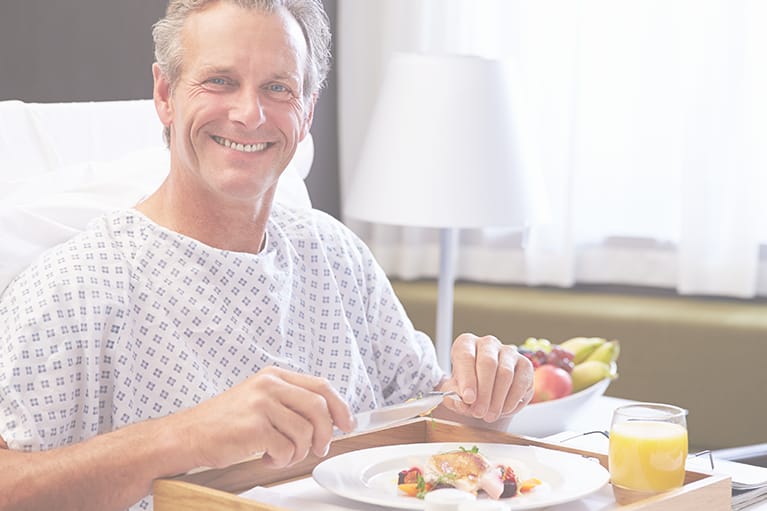 male patient is wearing a hospital gown, eating a meal in bed. The hospital uses nutrition services software.