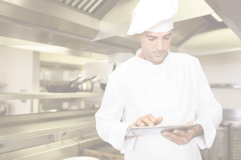 A male chef wearing a puffy hat looks down as he uses a tablet. He is standing in an industrial hospital kitchen automated by foodservice management software.