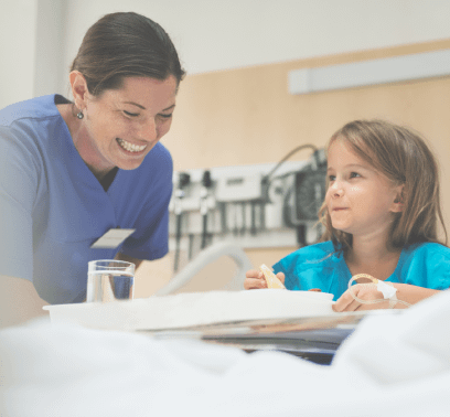 A female nurse grins as she sits by a young patient eating in a hospital bed.