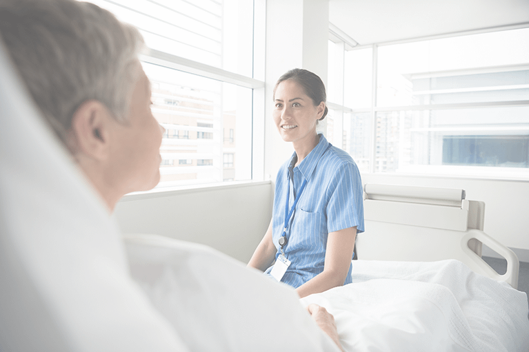 A female nurse smiles as she speaks with a hospital patient in a brightly lit room as they do patient bedside ordering.