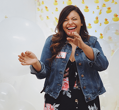 Jen Higgins is wearing a floral dress and denim jacket and laughing in front of a rubber ducky backdrop.