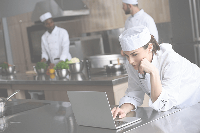 A female chef rests her chin on her hand as she looks at a laptop using automated hospital menu software in the kitchen.
