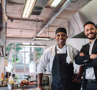 A chef and a professionally dressed man smile for the camera as they stand in a kitchen.