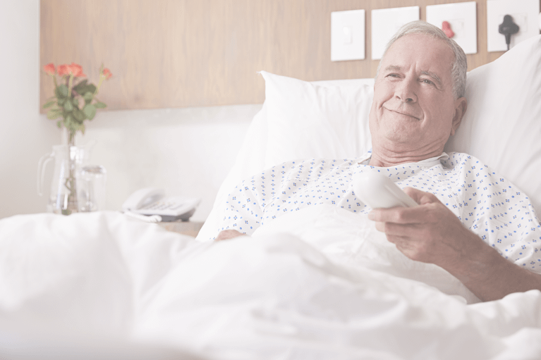 An elderly male patient using a remote for self ordering as he smiles and relaxes in his hospital bed.